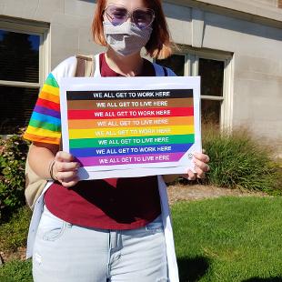 A student holding a rainbow flag sign that says "We all get to work here" and "We all get to live here"
