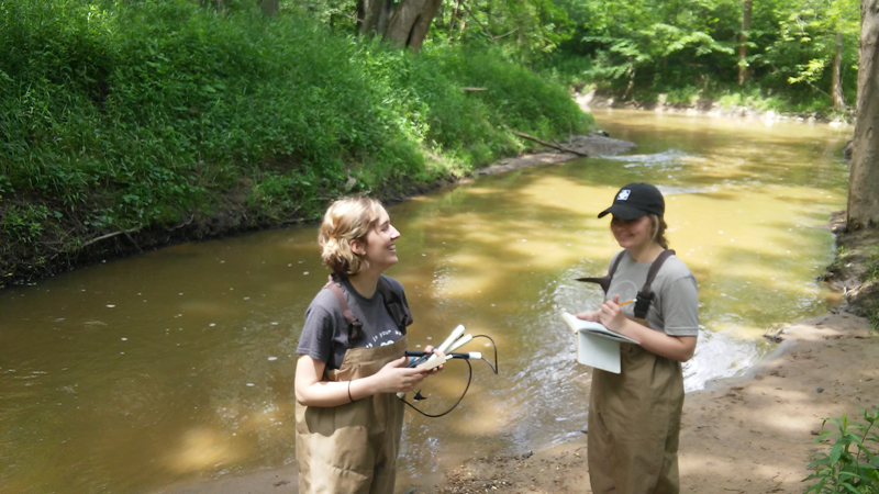 two students with stream flow measuring equipment in waders in a streambed